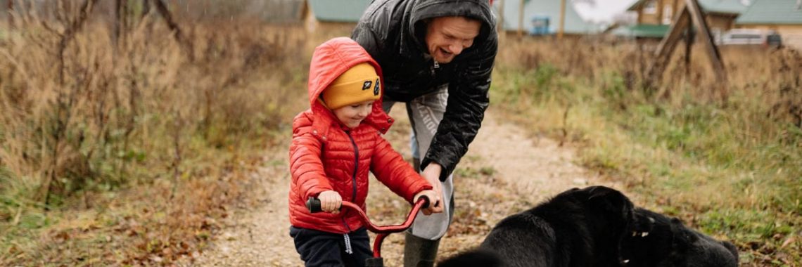 Dad helps son ride a bike on a path with family and dog, capturing a joyful outdoor moment.